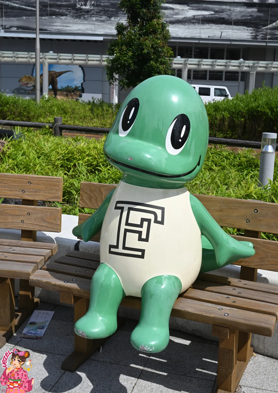 Mascotte verte de Fukui orn&eacute;e d'un F, assise sur un banc devant la gare