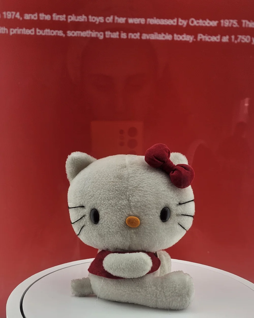 La peluche Hello Kitty d'origine de 1975, assise sur un socle blanc devant le panneau rouge de l'exposition