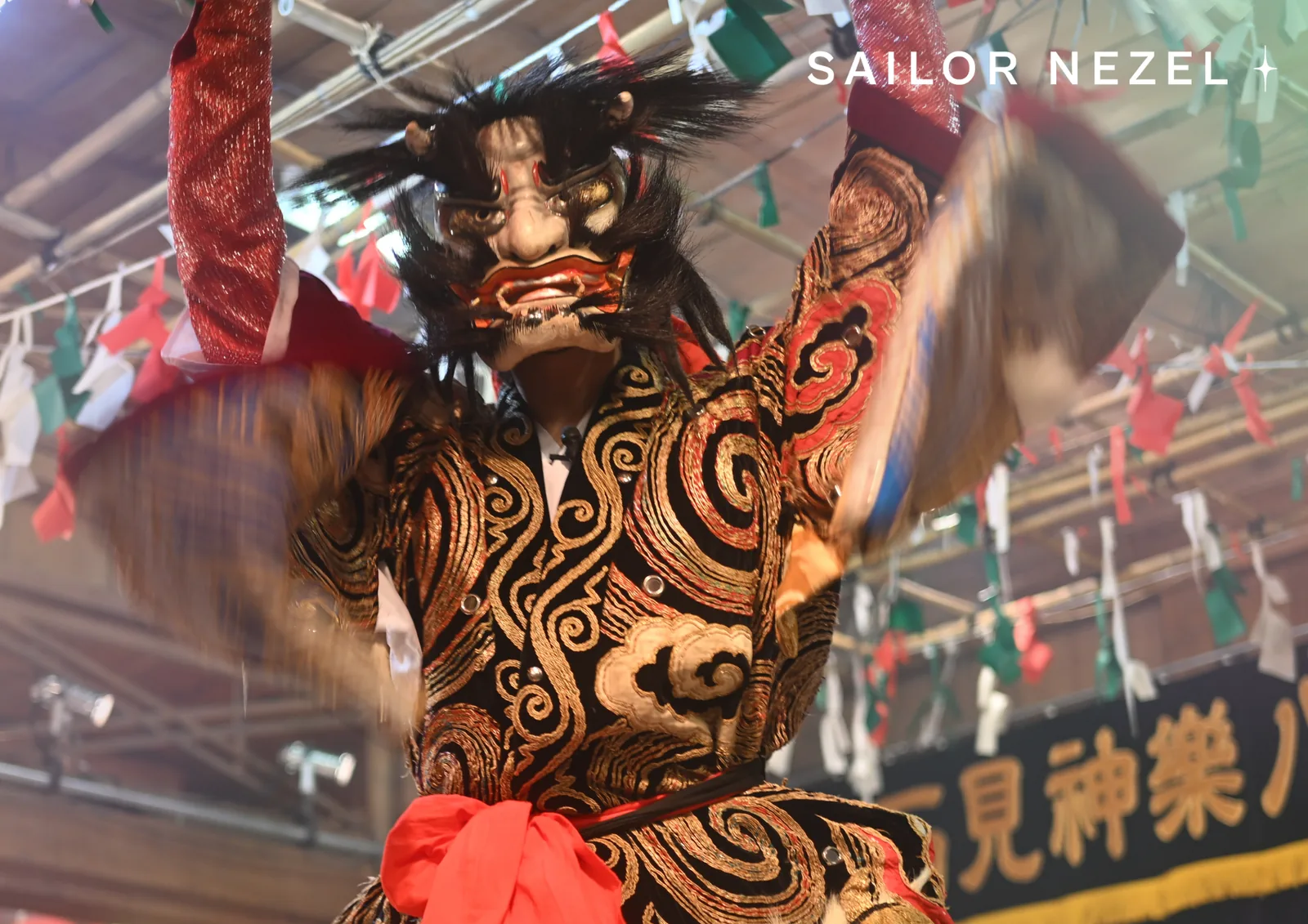 Danseur masqué en démon, bras levés et manches en mouvement, sous les banderoles de papier coloré du sanctuaire