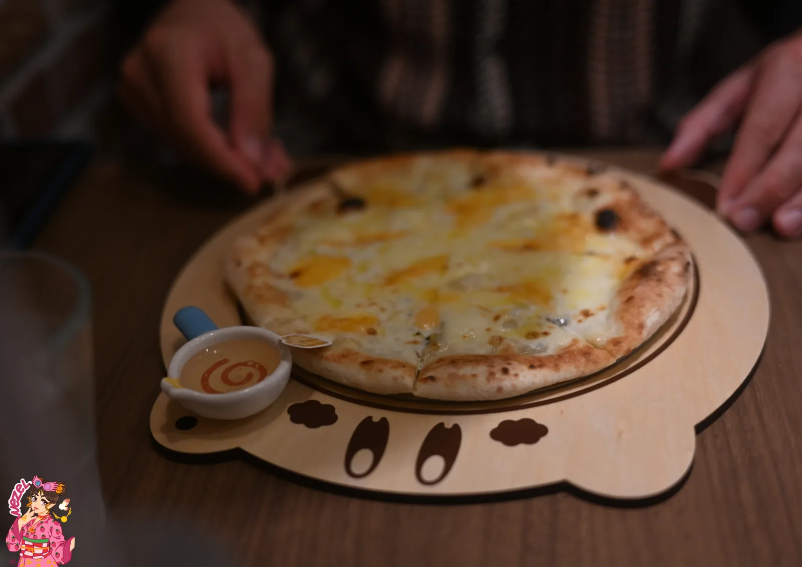 Pizza aux fromages posée sur un dessous de plat en bois en forme de Kirby, accompagnée d'un ramequin de sauce