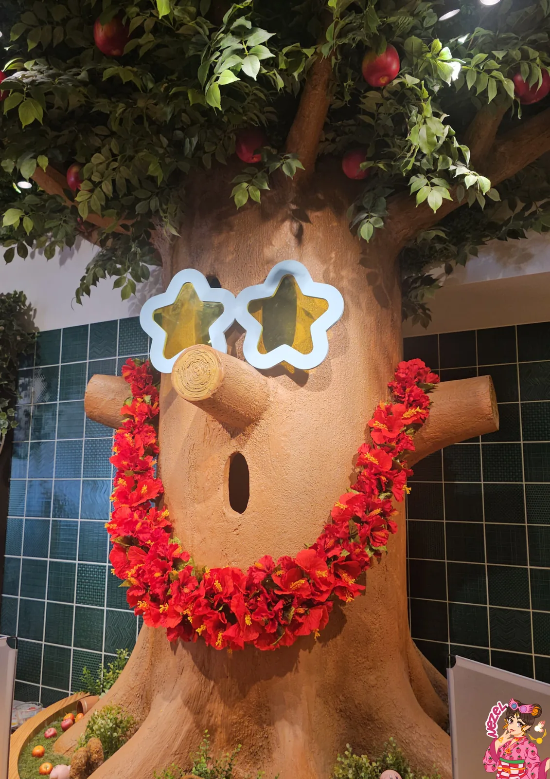 Whispy Wood, l'arbre géant du Kirby Café, avec ses lunettes en forme d'étoiles, son collier de fleurs rouges et ses pommes dans le feuillage