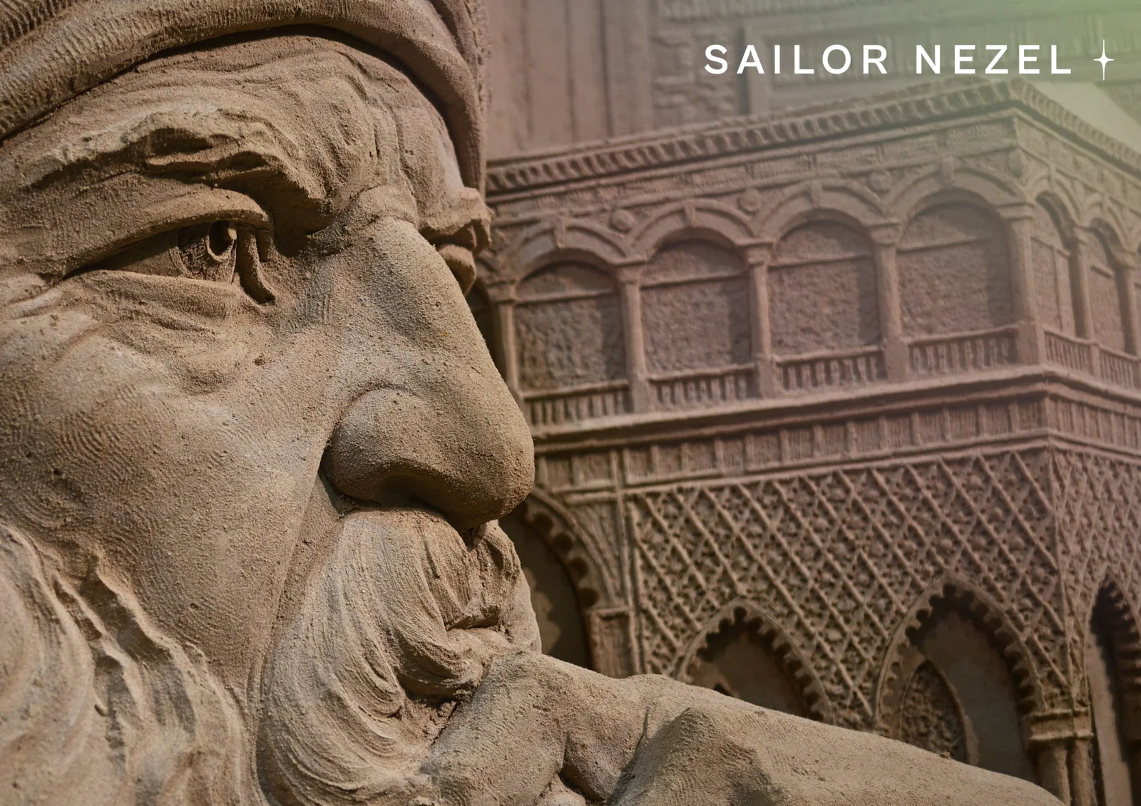 Gros plan sur un visage barbu coiffé d'un turban, sculpté dans le sable, devant un décor d'arcades et de dentelle de pierre