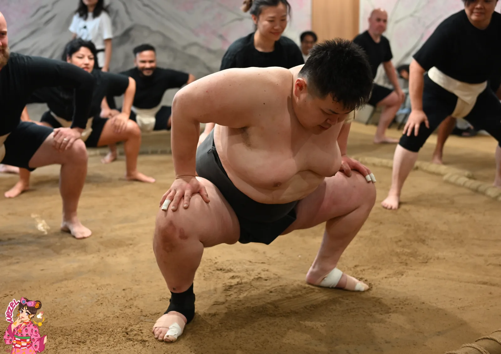 Des participants reproduisent la posture du shiko autour d'un rikishi, sur le dohyo